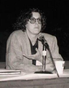 Fotografía en blanco y negro de Brenda Laurel durante una conferencia en los años 80, sentada frente a un micrófono, con gafas grandes, chaqueta de rayas y gesto expresivo mientras habla.